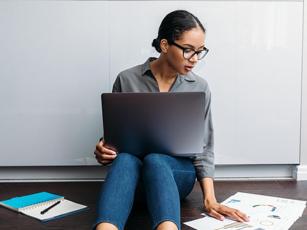 Femme assise sur le sol, travaillant avec un ordinateur portable sur les genoux. Des documents de travail sont éparpillés autour d'elle